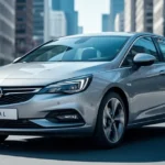 Srebrny Opel Astra L widziany z przodu i z boku na tle rozmytej miejskiej ulicy w słoneczny dzień.