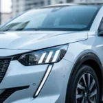 Srebrny Peugeot 208 II widziany z przodu i z boku, ukazujący nowoczesne reflektory LED na tle rozmytego miasta.