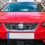 Czerwony Seat Ibiza V widziany od przodu, zbliżenie na grill i reflektory na tle rozmytej ulicy miejskiej.
