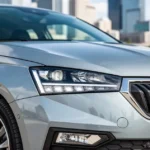 Nowoczesna Skoda Fabia IV w kolorze metalicznym, widoczna z przodu i z boku na tle rozmytego krajobrazu miejskiego.