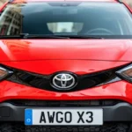 Jaskrawoczerwona Toyota Aygo X na tle rozmytej ulicy miejskiej, widok z przodu na nowoczesne reflektory i detale karoserii.