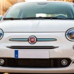 Przód białego Fiata 500 Hybrid z nowoczesnymi reflektorami zaparkowanego na słonecznej ulicy.