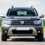Metaliczny szary SUV Dacia Duster II zaparkowany na asfaltowej drodze na tle rozmytego zielonego krajobrazu.