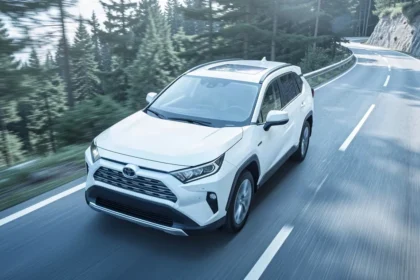 Biała Toyota RAV4 jadąca asfaltową drogą wśród gór i lasu w świetle dziennym.