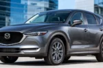 Metalicznie szara Mazda CX-5 II widziana z przodu pod kątem na tle nowoczesnych szklanych budynków w słoneczny dzień.