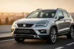 Srebrny Seat Ateca zaparkowany na asfaltowej drodze w promieniach zachodzącego słońca na tle nowoczesnego miasta.
