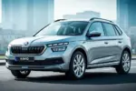 Srebrna Skoda Kamiq zaparkowana na asfaltowej drodze na tle rozmytego krajobrazu miejskiego.